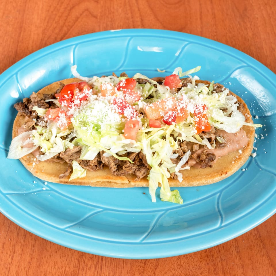 A large Mexican huarache with meat, beans, lettuce, tomatoes, and cheese on a blue plate.