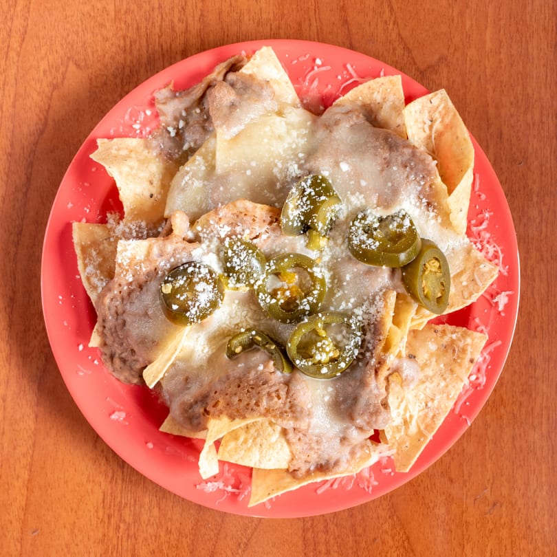 Nachos with melted cheese, refried beans, pickled jalapeños, and grated cheese on a red plate.