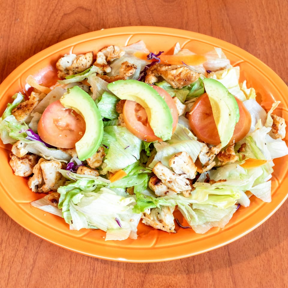 Grilled chicken salad with lettuce, tomatoes, avocado slices, and shredded carrots on an orange plate.