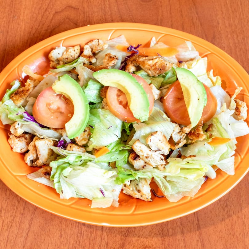 Grilled chicken salad with lettuce, tomatoes, avocado slices, and shredded carrots on an orange plate.