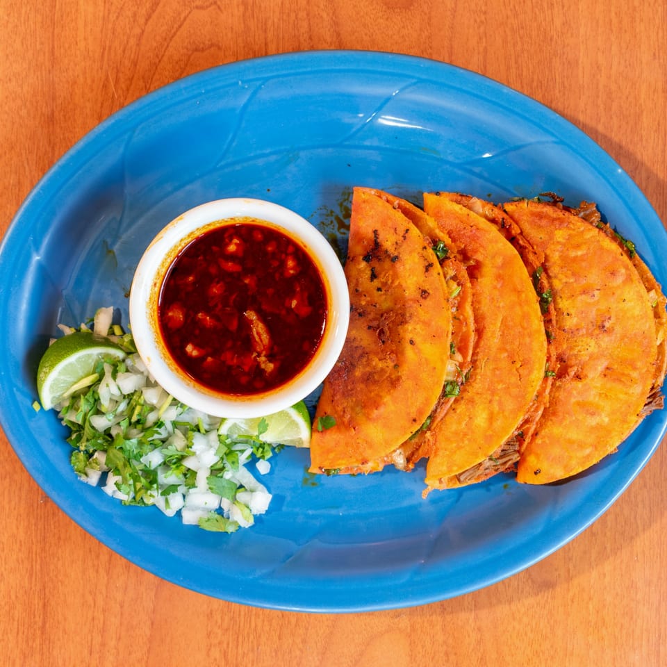 Four birria tacos with a side of consommé, chopped onions, cilantro, and lime wedges on a blue plate.