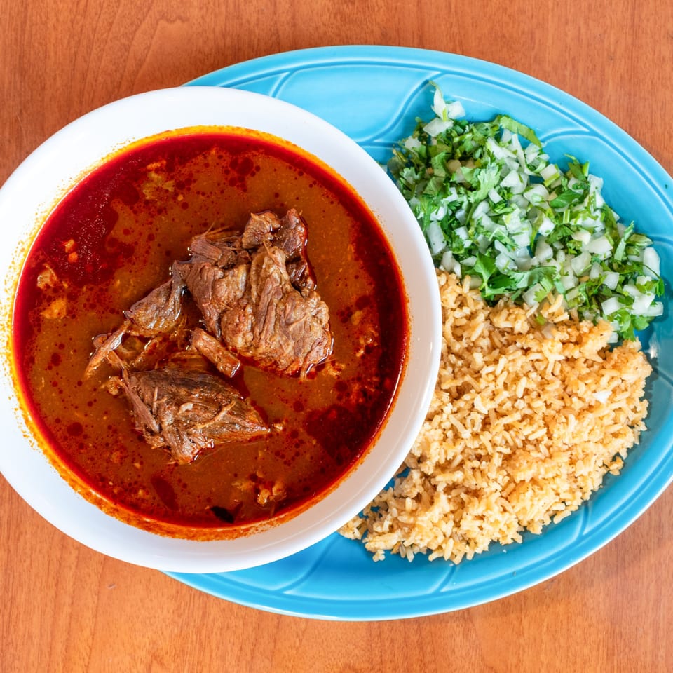 Hearty bowl of birria with rice, chopped cilantro, and onions on a wooden table.