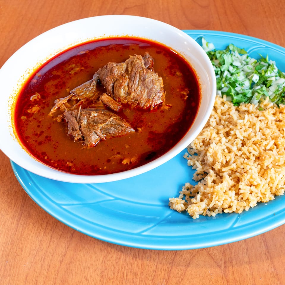 A bowl of birria with rice, chopped onions, and cilantro on a blue plate.