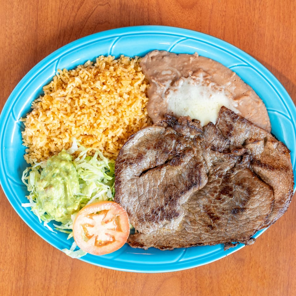 Grilled steak with rice, refried beans, guacamole, shredded lettuce, and a tomato slice on a blue plate.